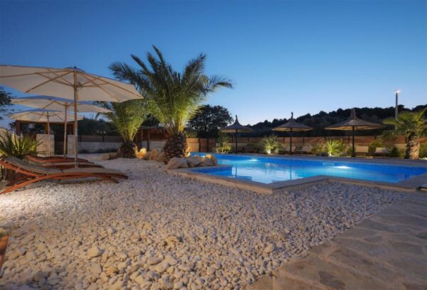 View of illuminated pool surrounded by rock covered terrace with sun loungers in sunset
